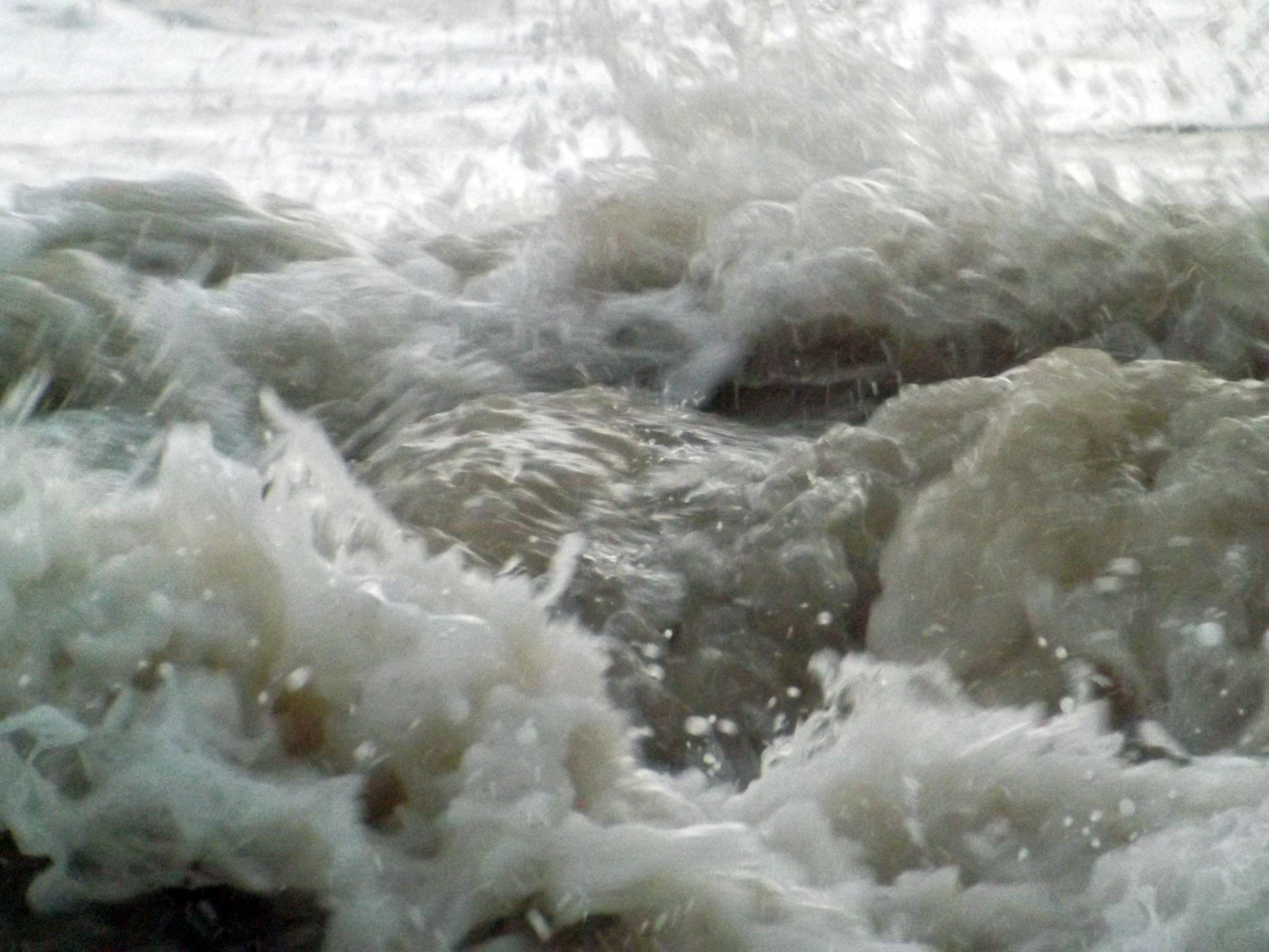 Close up of turbulent, foamy water crashing towards the camera. Grey, white, and light green tones.