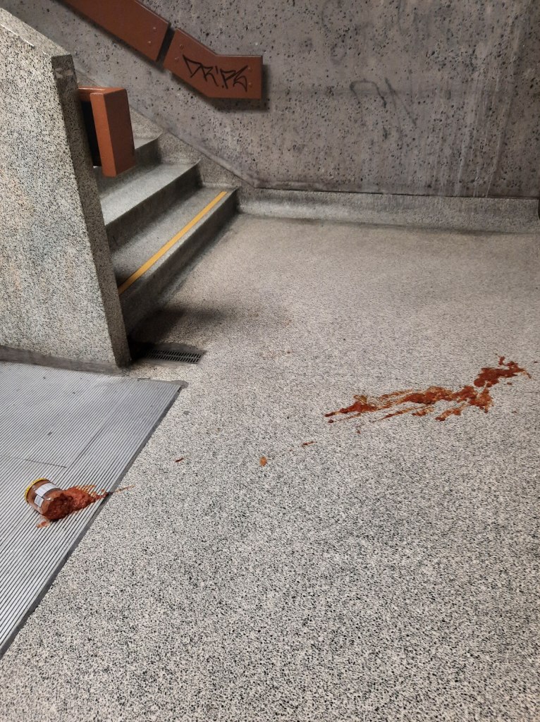 Photo of concrete ground and wall, with the bottom of a staircase in the top left part of the image. There is a smashed salsa jar on the ground that has splattered across the concrete.