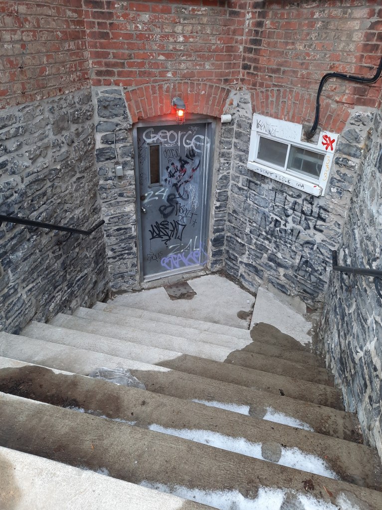 Photo of concrete stairs with melting snow on them leading down to a metal door with a small vertical window in it, which has a red light on above it and a window with a white frame to its right. The window and door are set into a brick building with grey bricks at the bottom and red bricks above. The door, window, and bricks are also covered in graffiti and tags. Additionally, there are two black handrails on either side of the stairs.