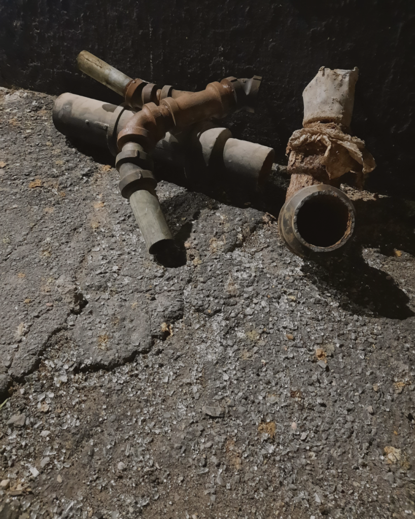 Four pieces of rusty pipe in a pile against a dark wall and on concrete, which is covered in a smattering of broken glass.